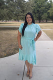 Woman in a light blue dress standing on a sidewalk with trees in the background
