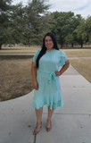 Woman in a light blue dress standing on a sidewalk with trees in the background