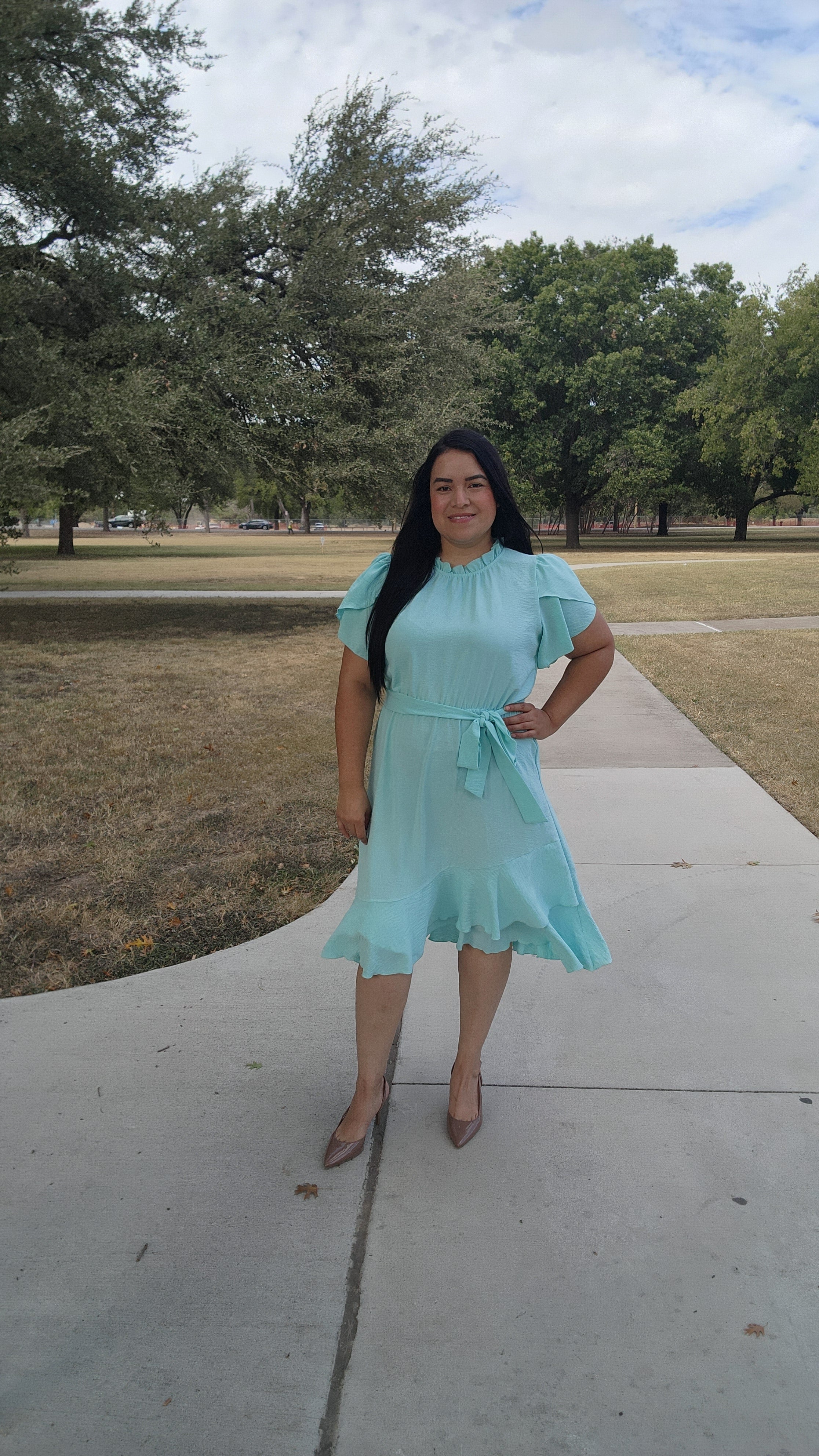 Woman in a light blue dress standing on a sidewalk with trees and grass in the background