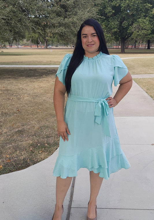 Woman wearing a light blue dress standing on a sidewalk with trees in the background