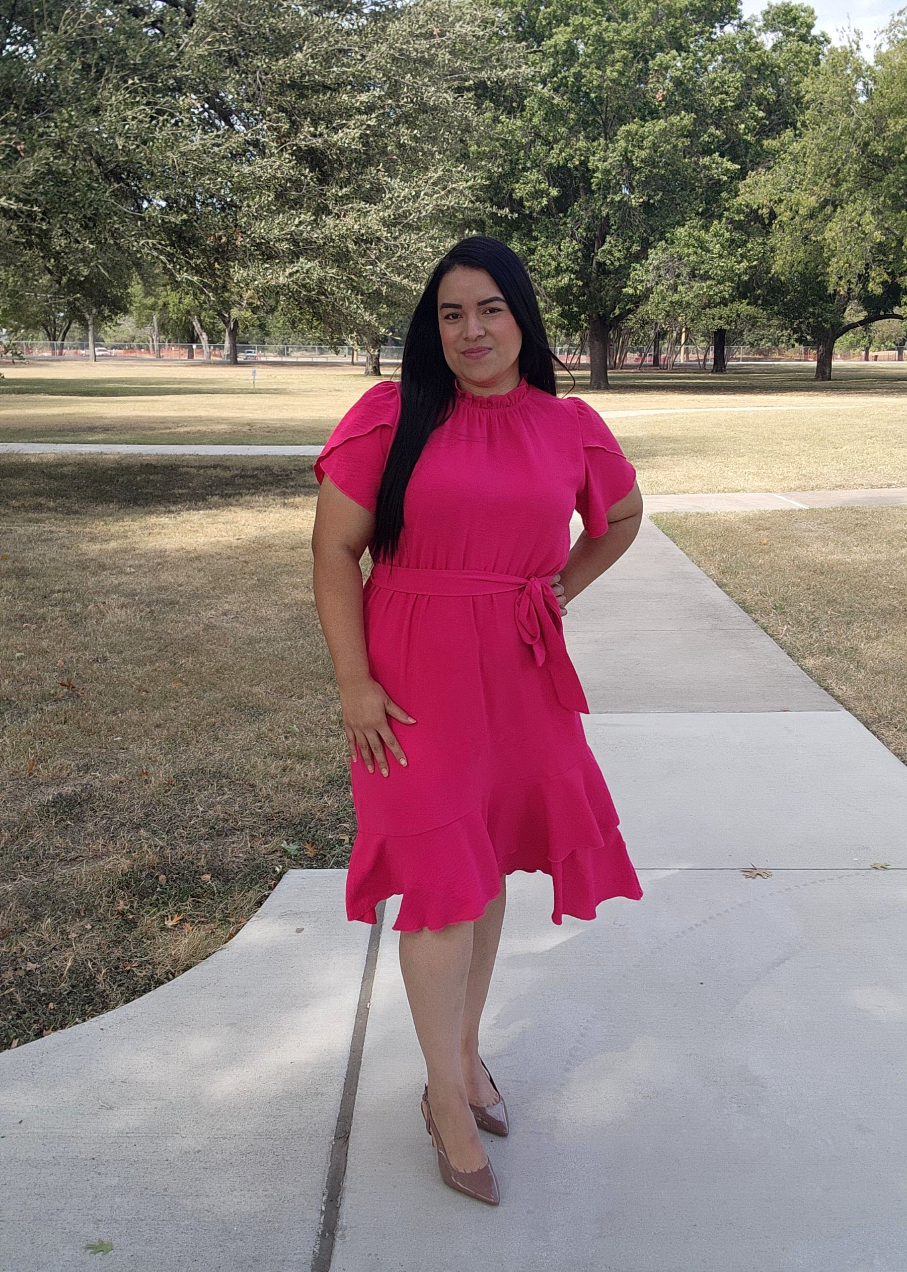Woman in a pink dress standing on a sidewalk with trees in the background