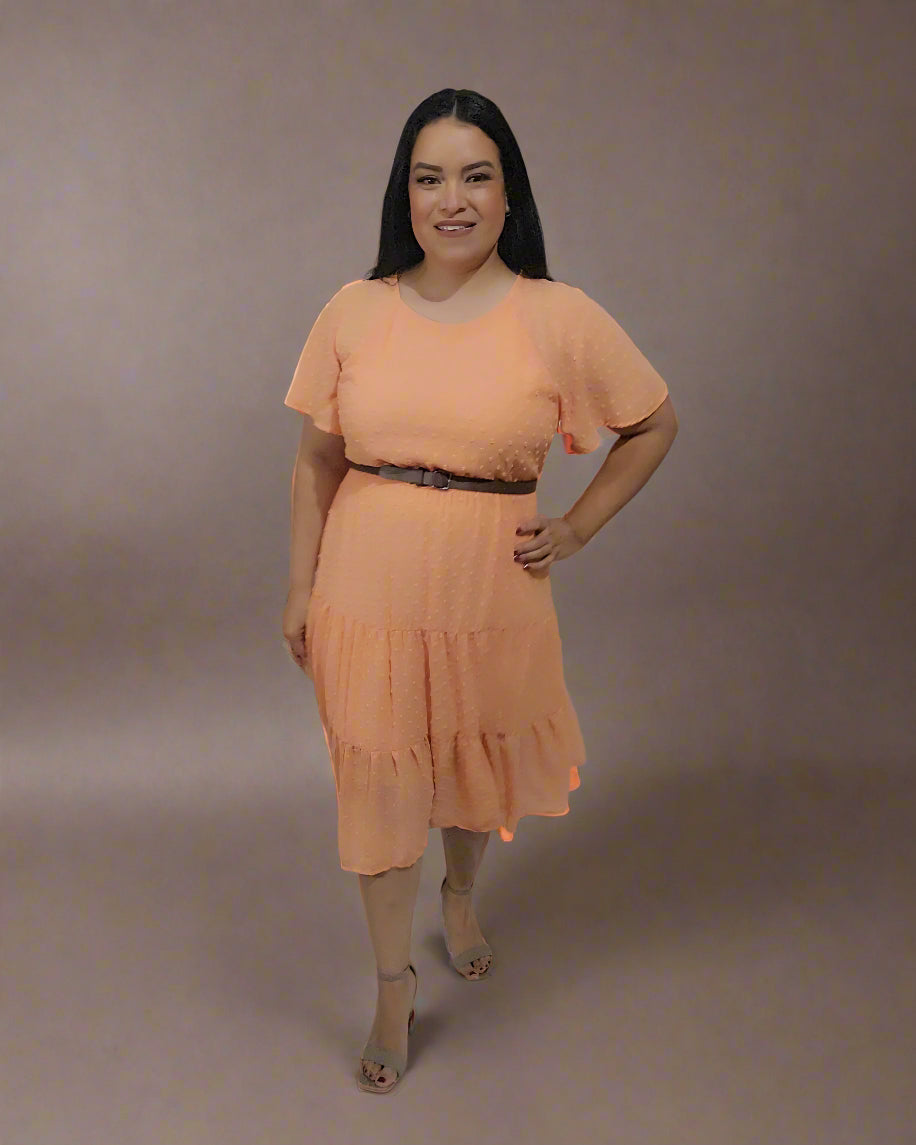Woman wearing an orange dress standing next to a small table with flowers against a white curtain background.