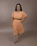 Woman wearing an orange dress standing next to a small table with flowers against a white curtain background.