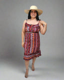 Woman wearing a colorful dress and wide-brimmed hat indoors with a white curtain background.