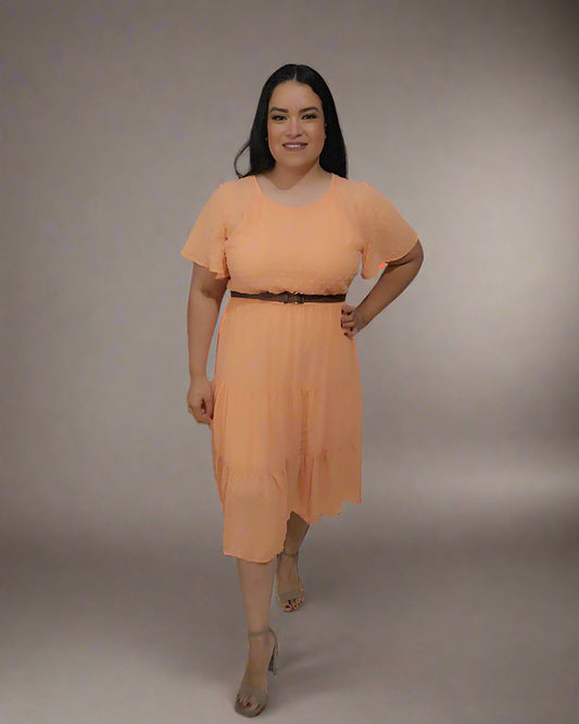Woman in an orange dress standing in a room with a white curtain and a small table with flowers.
