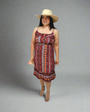 Woman in a patterned dress and wide-brimmed hat standing next to a small table with flowers against a white curtain background.